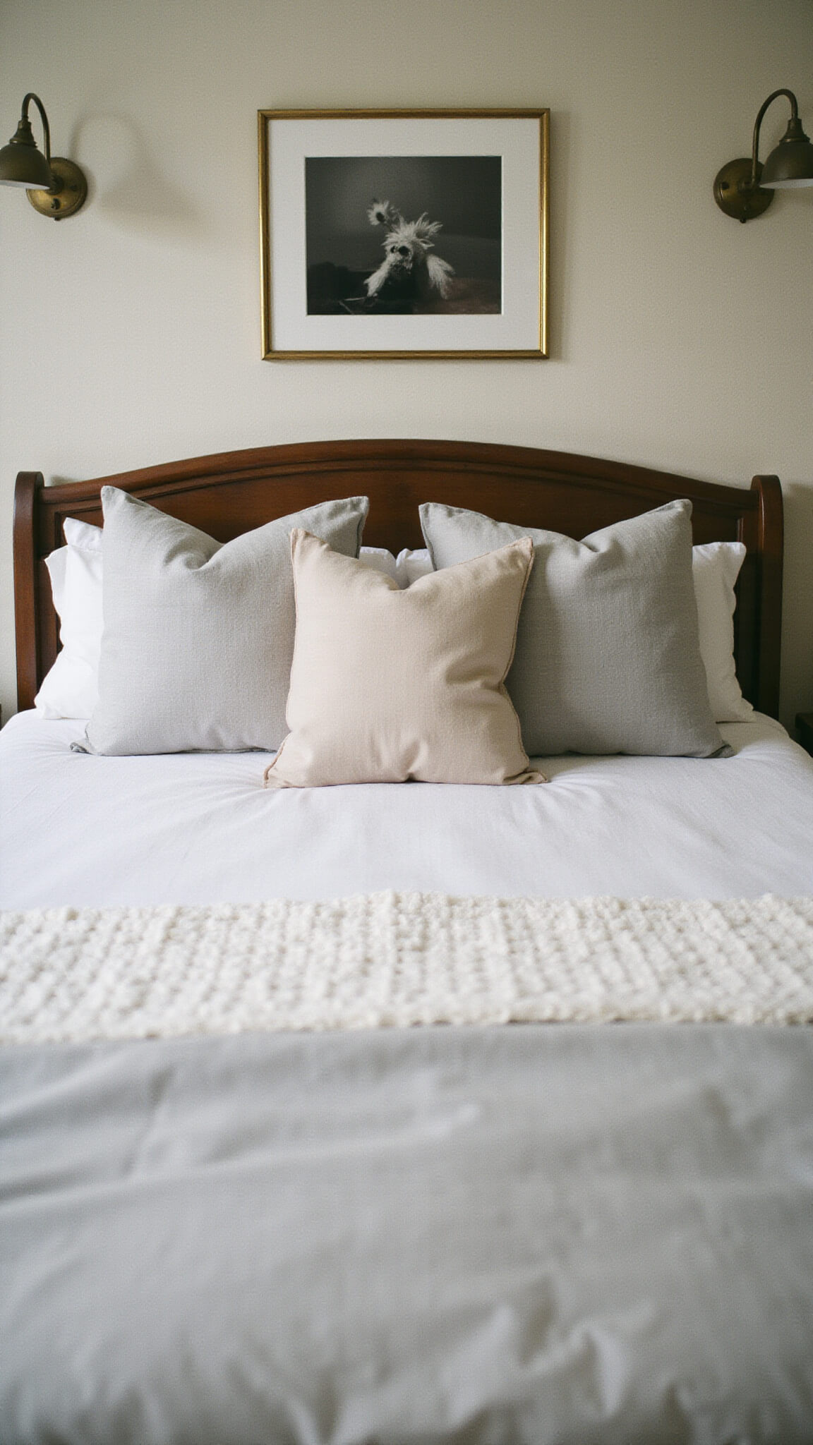 Elegant guest bedroom featuring layered white and pewter bedding with textured fabrics, warm brass lighting, and monochrome artwork above the bed.